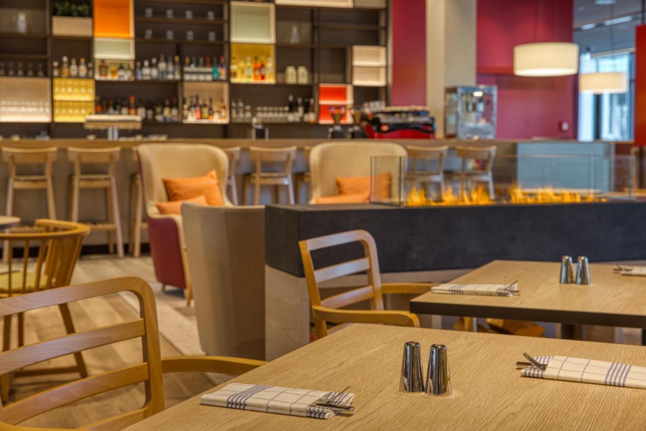 Intercityhotel Berlin Airport Ber Terminal 1 & 2 シェーネフェルト エクステリア 写真 The photo shows a modern restaurant or bar interior. In the foreground, there are wooden tables set with napkins and condiments. The seating includes wooden chairs and plush seating in various colors in the background. A stylish bar area is visible w